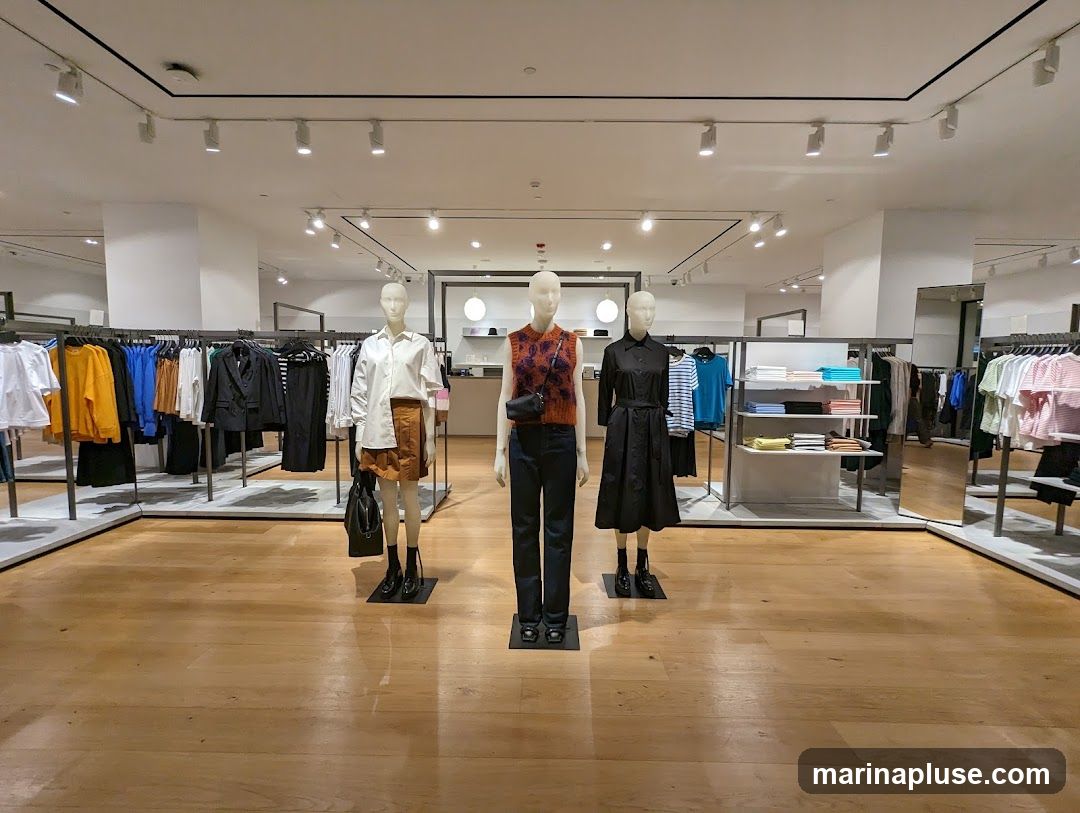 A well-lit clothing store interior featuring several mannequins displaying outfits. Racks of clothing are visible on the left and right sides of the image, with shelves of folded garments in the background. The floor is made of polished wood.