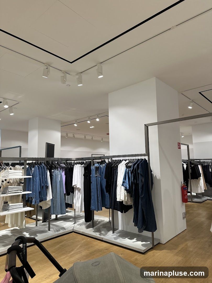 A view of the interior of a clothing store, with racks of clothes on display. The clothes are mostly in shades of blue, white, and black. There are also shelves with folded clothes and a stroller in the foreground. The store appears to be well-lit with track lighting on the ceiling.