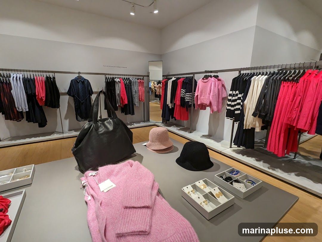 A well-lit clothing store interior featuring racks of women's apparel, including dresses, sweaters, and outerwear in various colors like pink, black, and red. A large black handbag and several hats are displayed on a table in the foreground, along with trays of jewelry. The store has a minimalist design with grey walls and wooden flooring.