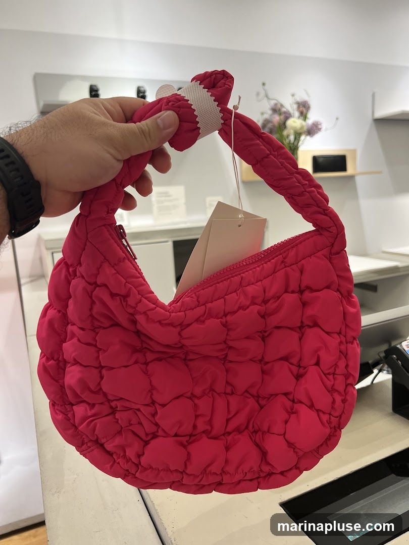 A hand holds a bright pink, puffy, quilted handbag with a braided handle. The bag has a zipper closure and a tag attached. In the background, there are shelves with items and a blurred floral arrangement.