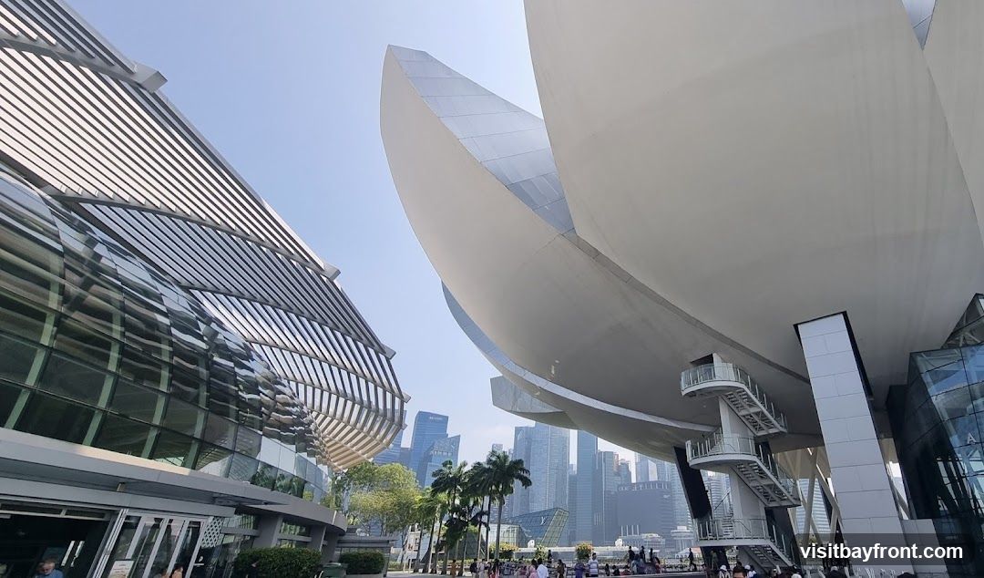 A low-angle shot showcasing the striking modern architecture of a building complex, with its distinctive curved roofline and glass facade. In the background, a dense city skyline with numerous skyscrapers is visible under a clear blue sky. Palm trees and a crowd of people suggest a public space or tourist attraction.