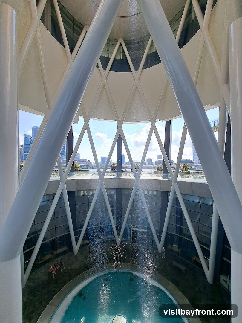 The image shows the interior of a modern building with a distinctive architectural design. Large white pillars and a white geometric ceiling structure dominate the foreground. Below, a circular pool with clear blue water is visible, with a gentle waterfall cascading into it. The background reveals a cityscape through large windows, suggesting the building is located in an urban area. A few people are seen sitting near the pool.