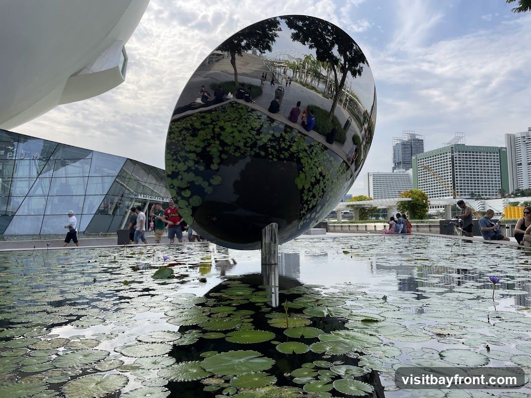 A large, reflective, oval-shaped sculpture stands in a pond filled with water lilies. The sculpture reflects the surrounding environment, including trees, people, and buildings. In the background, a modern building with a glass facade is visible, along with several high-rise buildings and a cloudy sky. People are seen sitting around the pond and walking near the building.