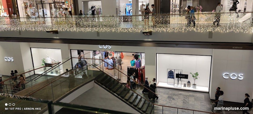 An interior view of a COS store in a mall, showing clothing displays and shoppers. The store features minimalist design with white walls and clothing racks filled with apparel. A prominent "SALE" sign is visible in one of the display windows. Escalators and railings are also visible, indicating the multi-level nature of the mall.