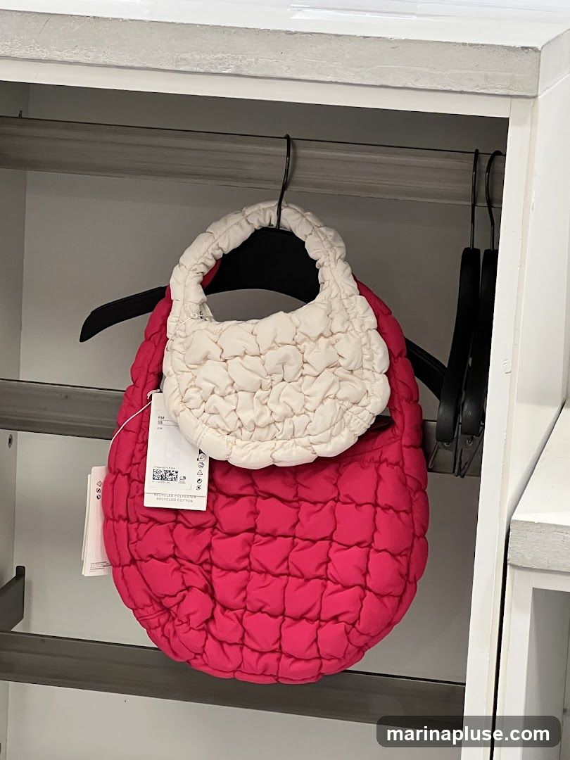 Two quilted bags, one cream and one bright pink, are displayed on hangers inside a white shelving unit. The pink bag is larger and hangs below the cream bag. Both bags have a ruched texture. A tag is visible on the pink bag.