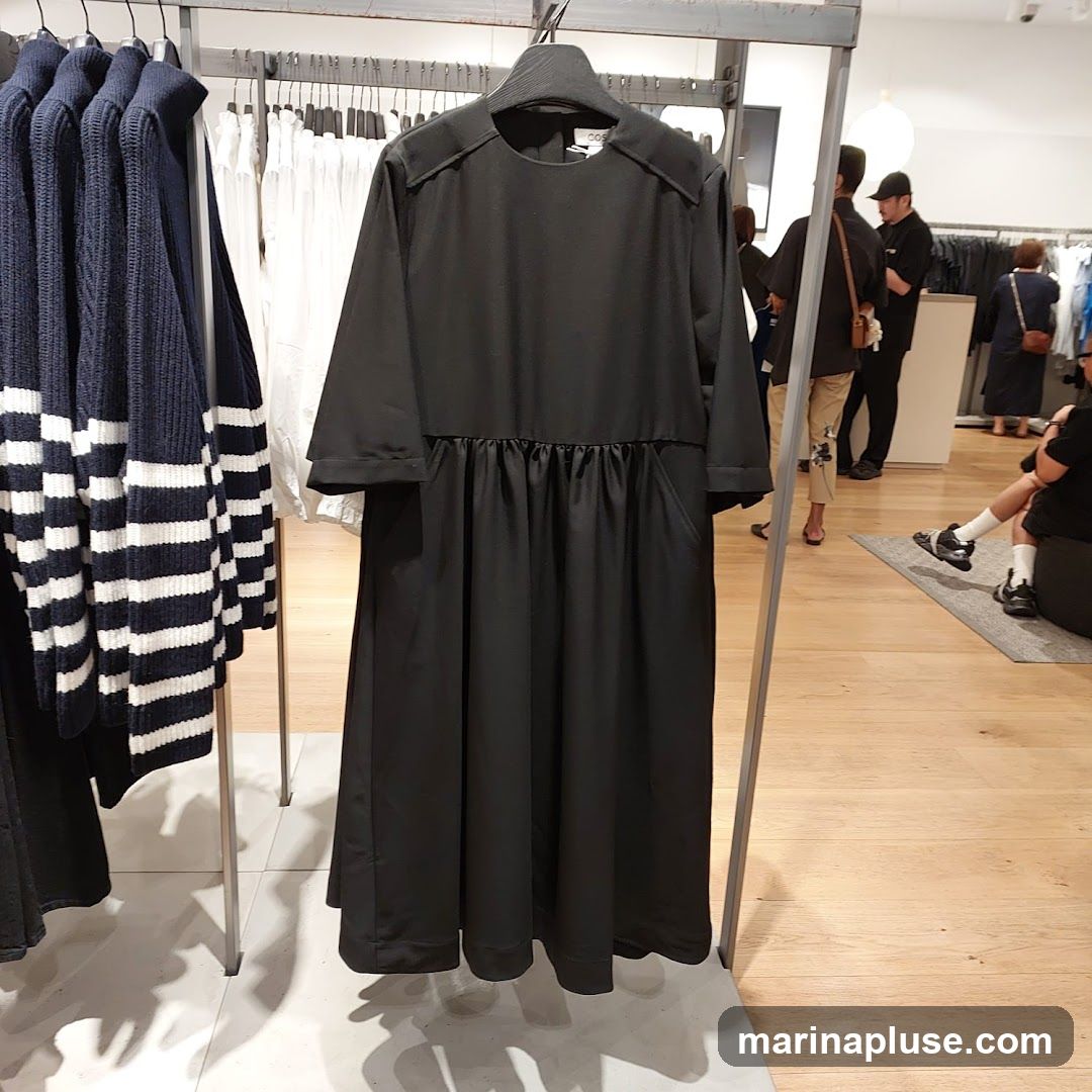 A black dress with a gathered waist and pockets is displayed on a hanger in a clothing store. To the left, several navy and white striped sweaters are also on display. In the background, other shoppers are browsing the merchandise.