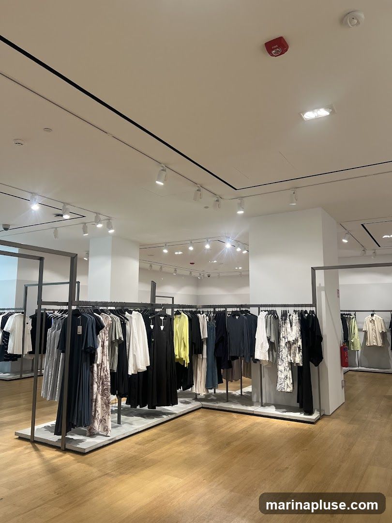 A well-lit clothing store interior with racks of women's apparel displayed on a light wooden floor. The clothing includes dresses, skirts, and tops in various colors and patterns. The store features modern track lighting and a clean, minimalist design.