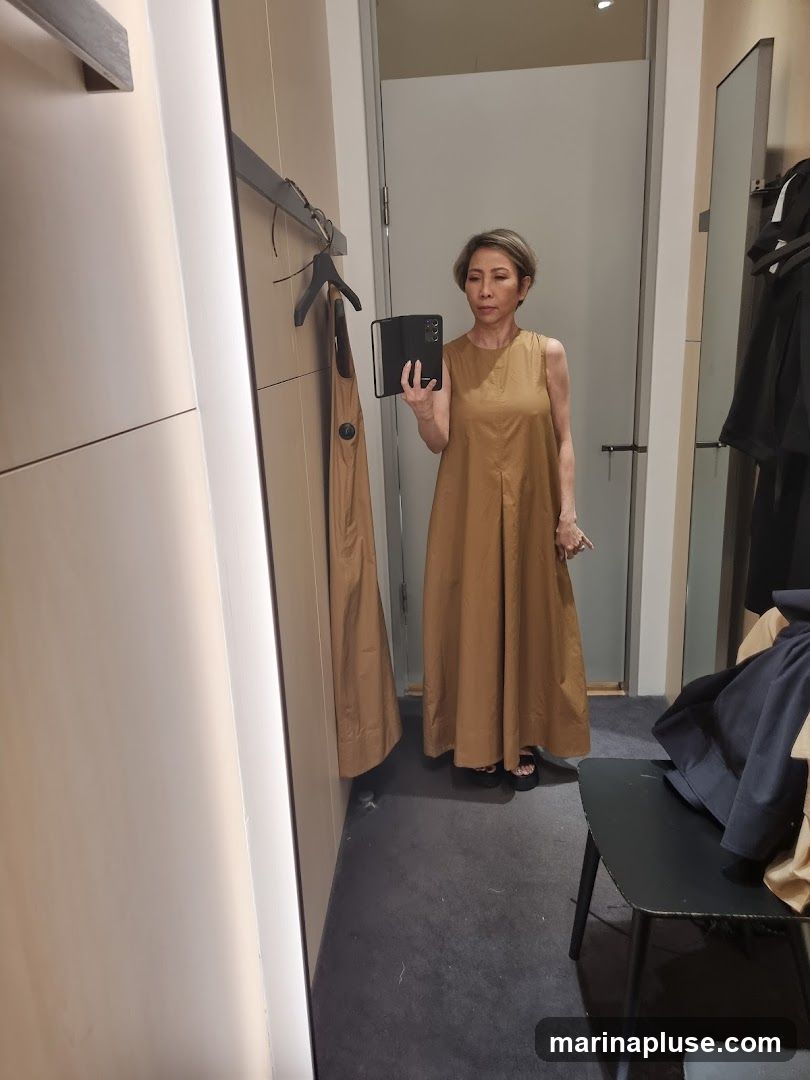 A woman with short, gray hair is standing in a fitting room, taking a mirror selfie. She is wearing a sleeveless, floor-length, tan-colored dress with pleats at the waist. The fitting room has a large mirror, a rack with clothes, and a stool.
