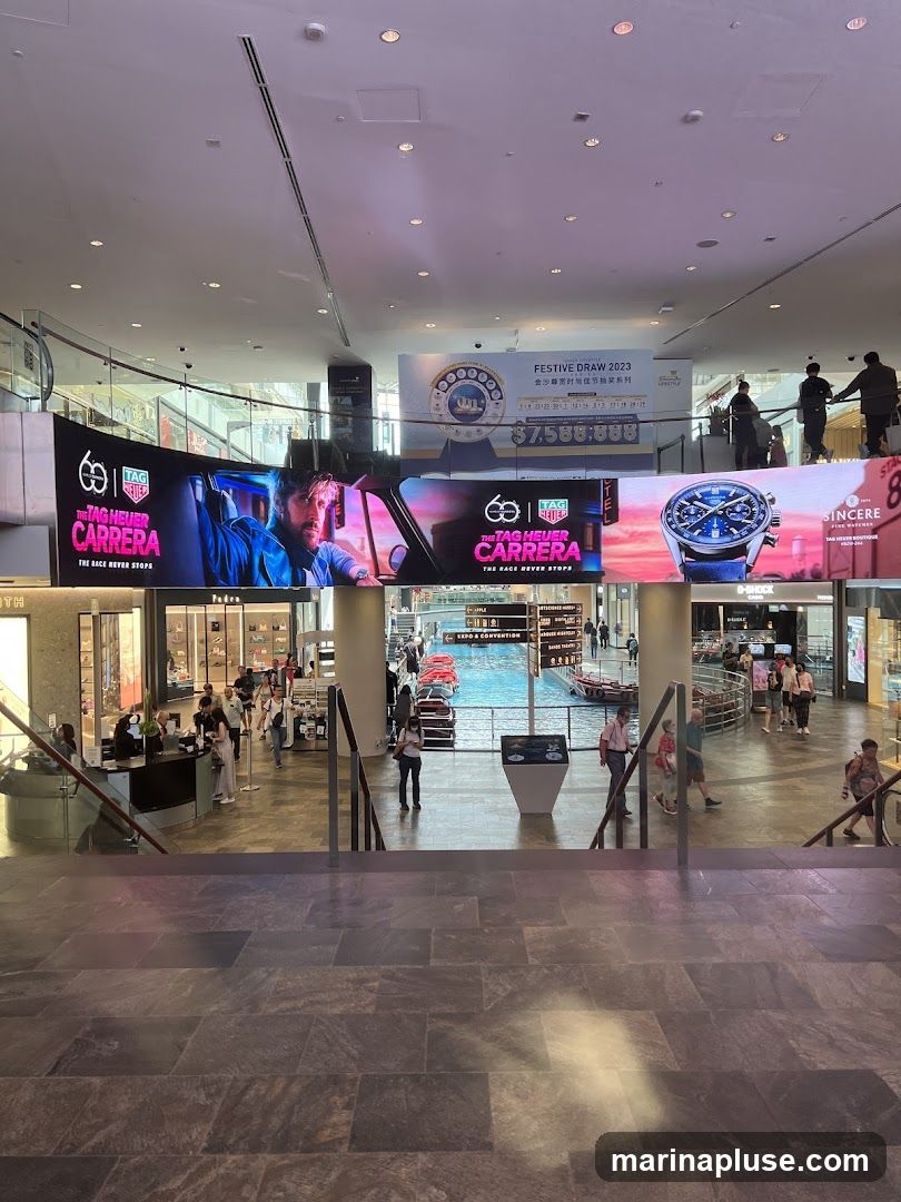A wide shot of the interior of a modern shopping mall. Large digital screens display advertisements for TAG Heuer Carrera watches and a "Festive Draw 2023" promotion with a prize of $7,588,888. People are seen walking around the mall, and shops are visible on the lower levels. In the background, a canal with boats can be seen.