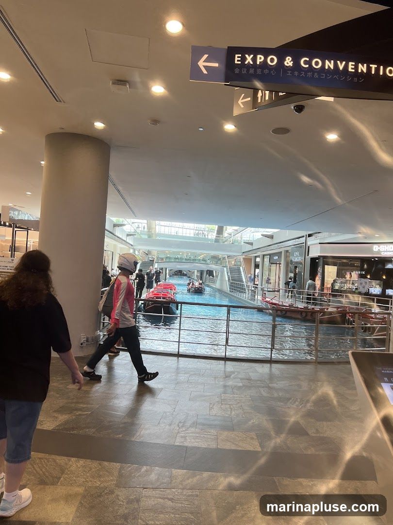 An indoor scene with a canal running through a shopping mall. Several small boats with passengers are on the canal. Shops are visible on the upper levels surrounding the canal. Signage indicates directions to an "EXPO & CONVENTION" area.