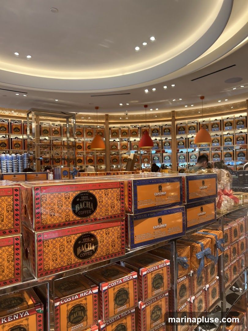 A display of numerous boxes of Bacha Coffee, featuring various origins like Kenya, Costa Rica, Yemen, and Explorer blends. The boxes are stacked on shelves and in display cases, with some wrapped in gift packaging. The store interior is visible in the background, with warm lighting and more coffee boxes lining the walls.