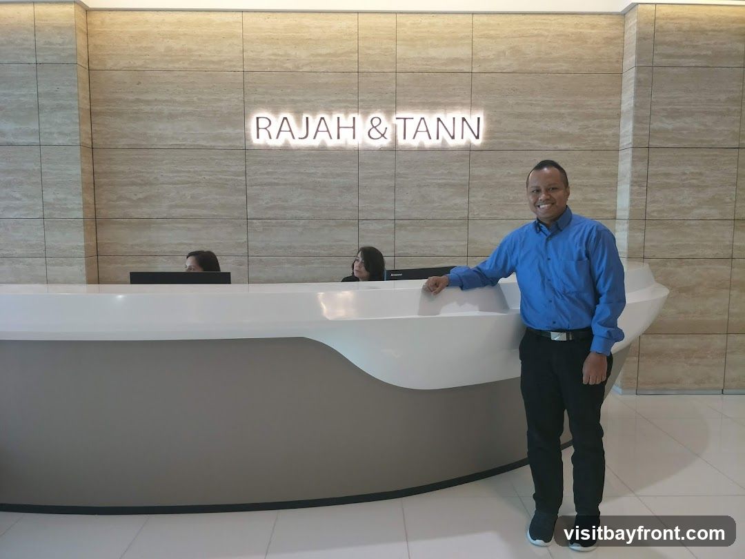 A man in a blue shirt and black pants stands at a reception desk in an office. Behind him, the words "RAJAH & TANN" are illuminated on a tiled wall. Two women are visible behind the reception desk, appearing to work there.