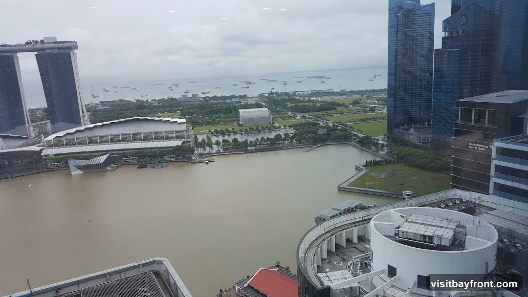An aerial view of a city skyline featuring the iconic Marina Bay Sands hotel and several modern skyscrapers. A body of water, likely a bay or river, is in the foreground, with numerous ships anchored in the distance. The scene is overcast, suggesting a cloudy day. The buildings are a mix of glass and steel, reflecting the muted light.