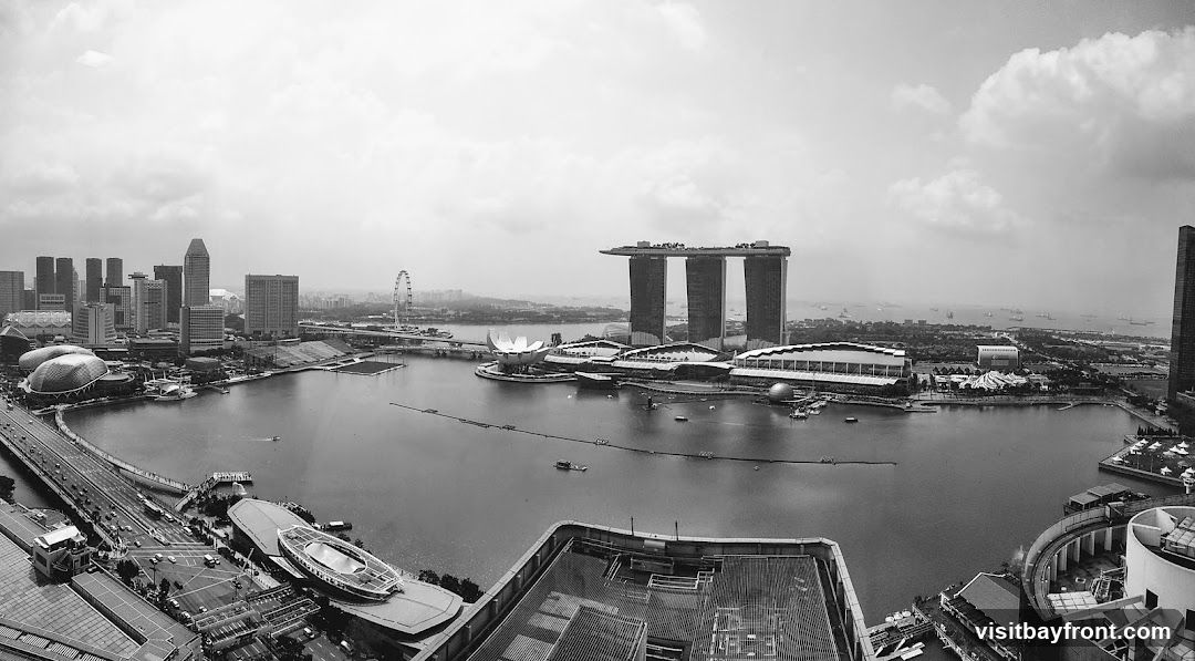 A black and white panoramic photograph captures the expansive Singapore skyline, featuring the iconic Marina Bay Sands hotel with its distinctive three towers and rooftop infinity pool. To the left, the ArtScience Museum, shaped like a lotus flower, is visible. The cityscape includes numerous skyscrapers, a Ferris wheel, and a bridge spanning across the water. The bay is dotted with boats and ships, and the horizon is hazy with clouds. The foreground shows rooftops of modern buildings.