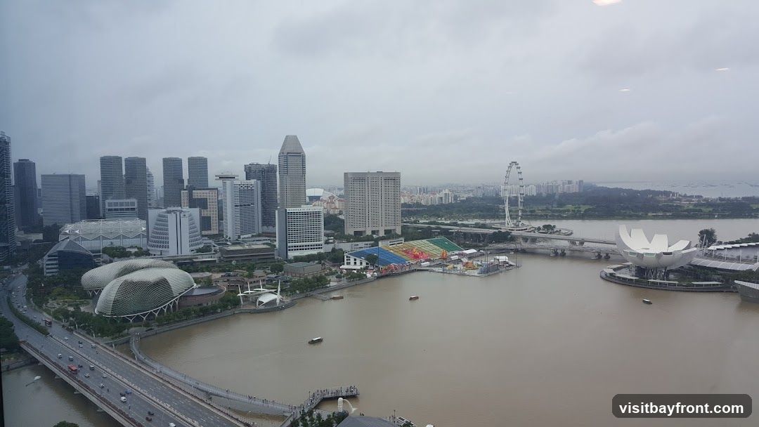 An aerial view of the Singapore skyline and Marina Bay on a cloudy day. The image features numerous skyscrapers, the Marina Bay Sands resort with its distinctive boat-shaped top, the ArtScience Museum with its lotus-like design, the Singapore Flyer Ferris wheel, and a bridge spanning the bay. The water in the bay appears murky. Several boats are visible on the water. In the foreground, a highway with traffic can be seen.