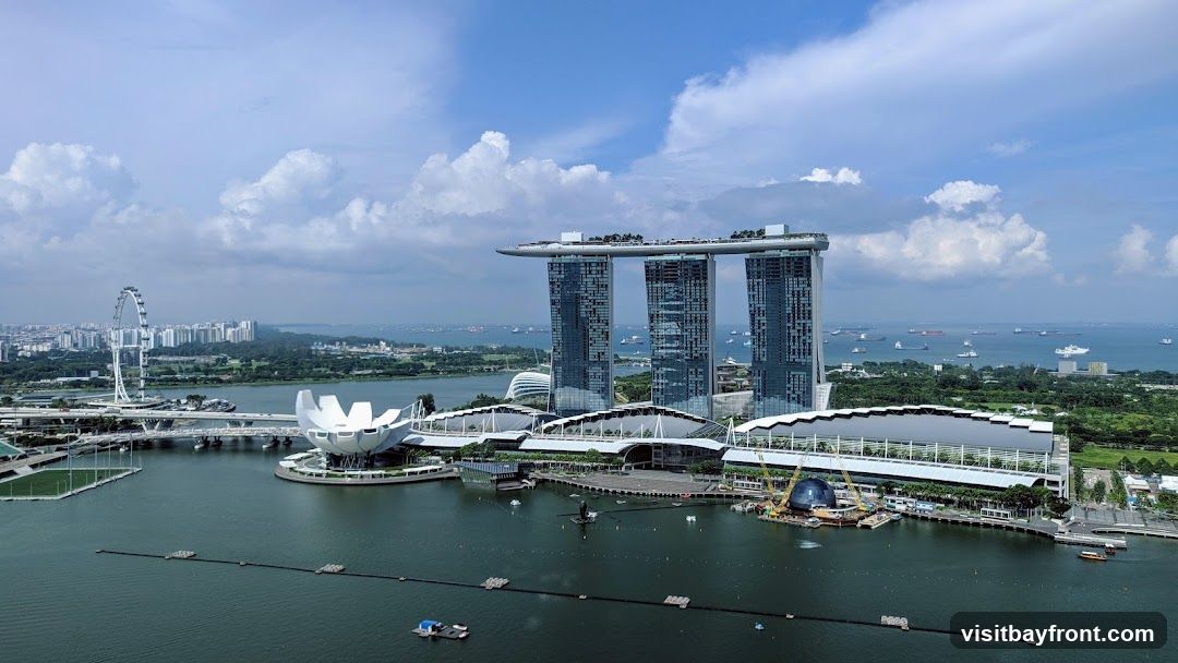 An aerial view of the Marina Bay Sands hotel and casino complex in Singapore, with its iconic three towers connected by a rooftop infinity pool. The ArtScience Museum, shaped like a lotus flower, is visible in the foreground. The Singapore Flyer Ferris wheel can be seen in the distance, along with a busy shipping lane filled with numerous cargo ships and tankers. The cityscape of Singapore stretches across the horizon under a partly cloudy sky.