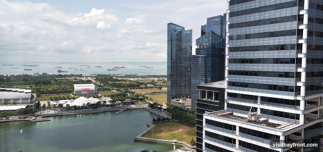 A panoramic view of a modern cityscape featuring numerous skyscrapers and a busy harbor filled with ships. In the foreground, a body of water reflects the sky and surrounding buildings. Beyond the water, there are green spaces and large, architecturally distinct buildings, including one with a prominent Singaporean flag. The background shows a vast expanse of the sea dotted with cargo ships under a cloudy sky.