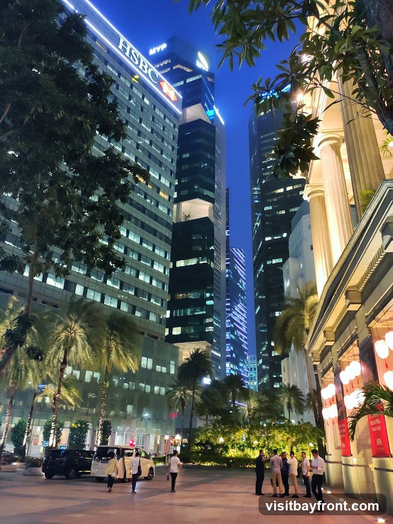 A nighttime view of a modern city with tall skyscrapers illuminated by lights. In the foreground, there are palm trees and a group of people standing on a paved area. The buildings feature contemporary architecture, with the HSBC logo visible on one of the prominent towers. The scene conveys a sense of urban sophistication and activity.
