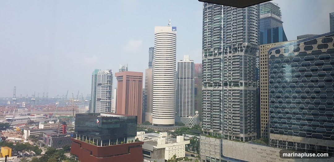 A panoramic view of a modern city skyline featuring numerous skyscrapers of varying architectural styles. In the foreground, several tall buildings dominate the scene, some with intricate facade designs. In the background, a sprawling port with many cranes is visible under a hazy sky.