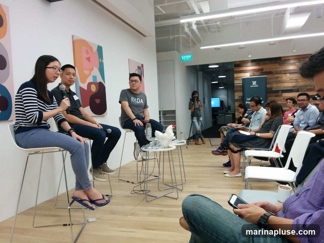 A panel discussion is taking place in a modern office space. Three people are seated on high stools on a stage, with microphones. The woman on the left is speaking. Behind them are colorful abstract paintings. An audience is seated in chairs facing the stage. A white chicken figurine is on a small table. In the background, a banner with the Zendesk logo is visible, along with an "EXIT" sign. A person in the foreground is looking at their phone.
