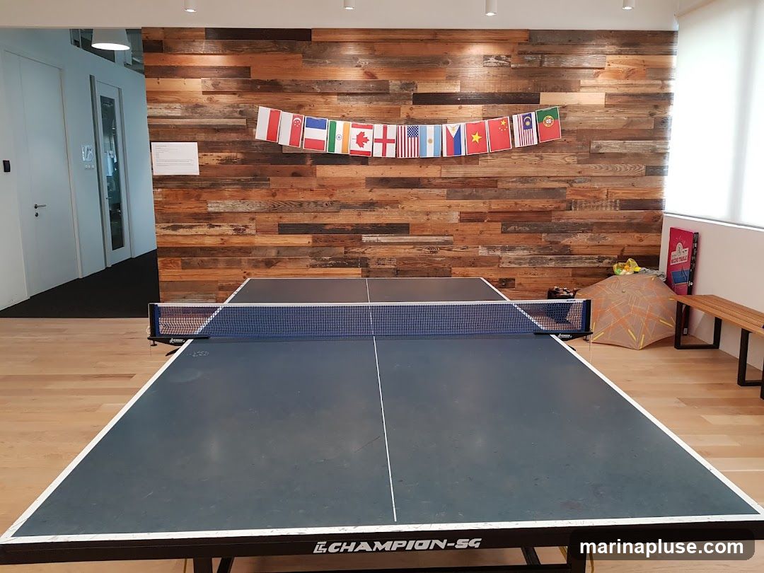 A ping pong table is set up in a break room with a rustic wooden accent wall. A string of international flags hangs above the table. To the right, there is a bench, an umbrella, and a sign that says "Nacho Table".