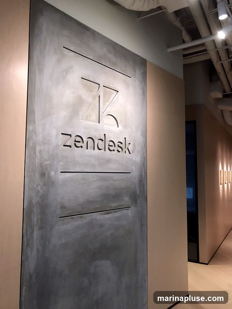A close-up view of a wall with the Zendesk logo and name prominently displayed. The logo is a stylized 'ZK' design, and the word 'Zendesk' is below it, both carved into a textured gray panel. The panel is framed by light wood-colored walls. The background shows a hallway with lighting fixtures on the wall and exposed ceiling pipes.