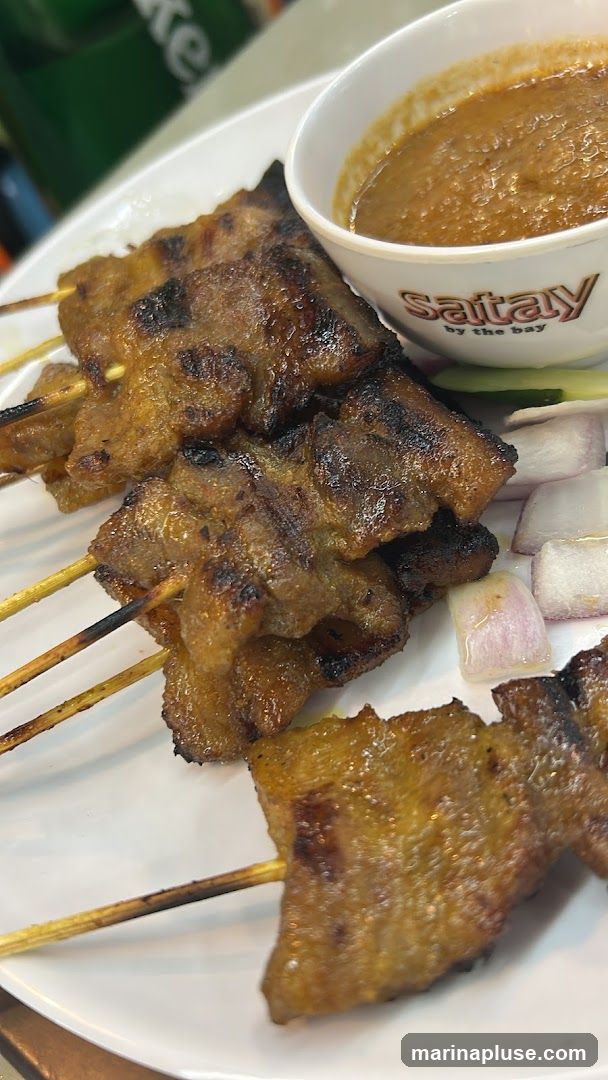 A close-up shot of grilled satay skewers served on a white plate. The skewers are accompanied by a bowl of peanut sauce, sliced red onions, and cucumber. The logo 'satay by the bay' is visible on the side of the sauce bowl.