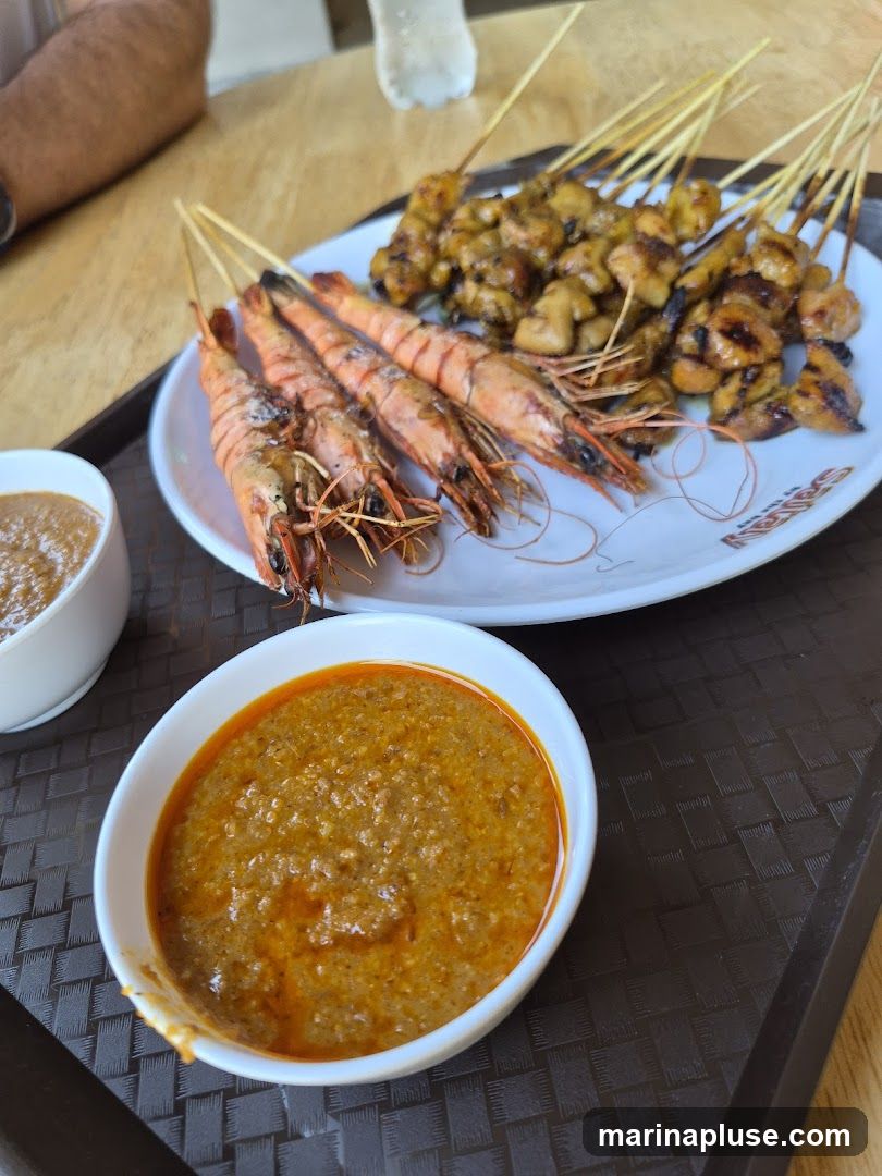 A close-up shot of a meal featuring grilled prawns and satay skewers served with peanut sauce. The prawns are arranged on a white plate alongside several skewers of grilled meat, likely chicken satay. Two bowls of peanut sauce are visible, one in the foreground and another slightly out of focus to the left. The food is presented on a dark, textured tray on a wooden table. A person's arm is visible in the upper left corner.