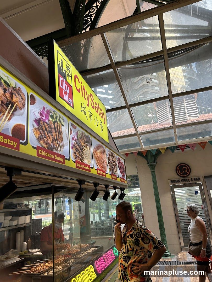 A food stall named City Satay displays images of various satay dishes. A man in a colorful shirt is looking at the food. In the background, there is an entrance to an Indian Grill & Bar.