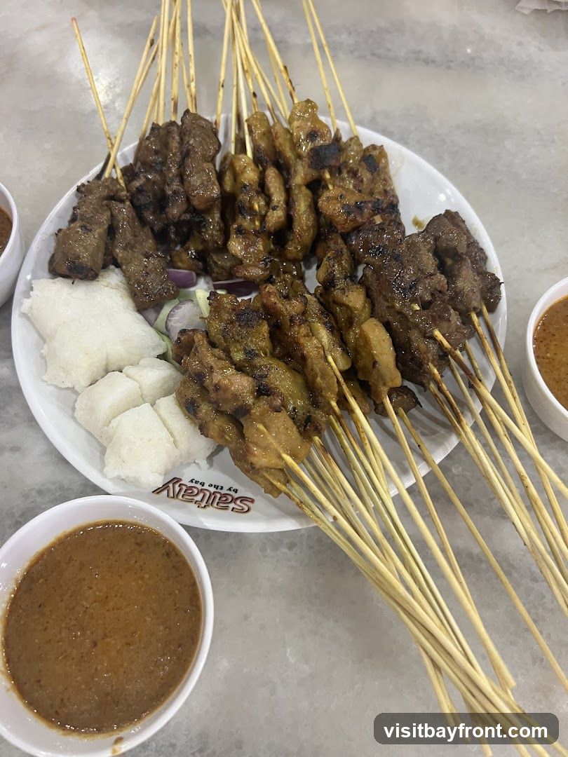 A white plate filled with numerous skewers of grilled satay, accompanied by cubes of white rice and bowls of peanut sauce. The plate has a logo that reads "Saitay by the bay".