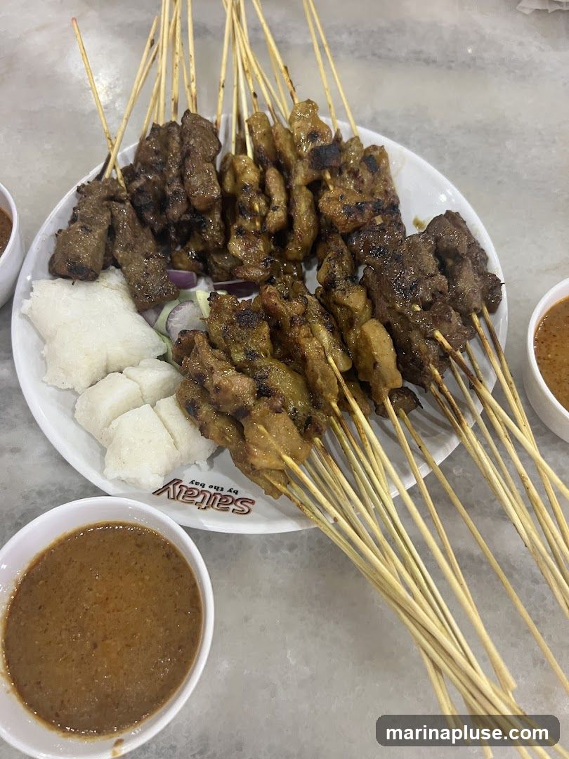A white plate filled with numerous skewers of grilled satay, accompanied by cubes of white rice and bowls of peanut sauce. The plate has a logo that reads "Saitay by the bay".
