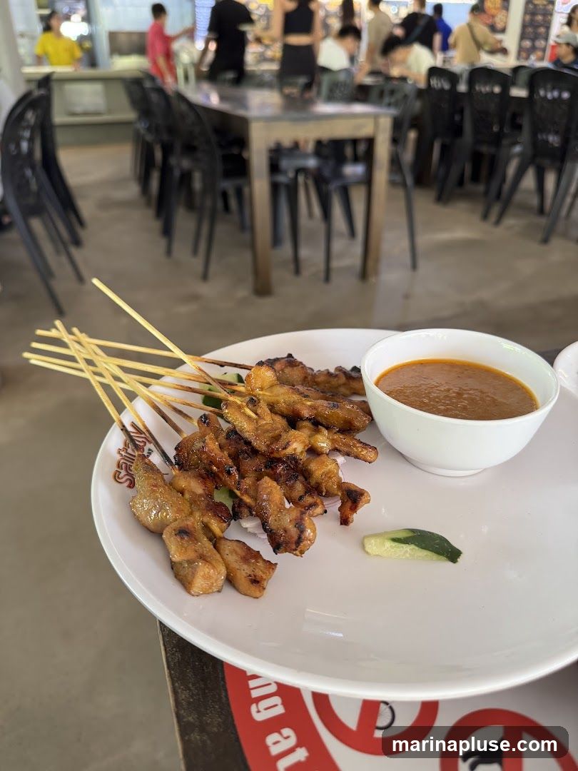A plate of grilled satay skewers served with a side of peanut sauce and cucumber slices. The skewers appear to be chicken or pork. In the background, a busy food court or hawker center is visible with tables, chairs, and people dining.
