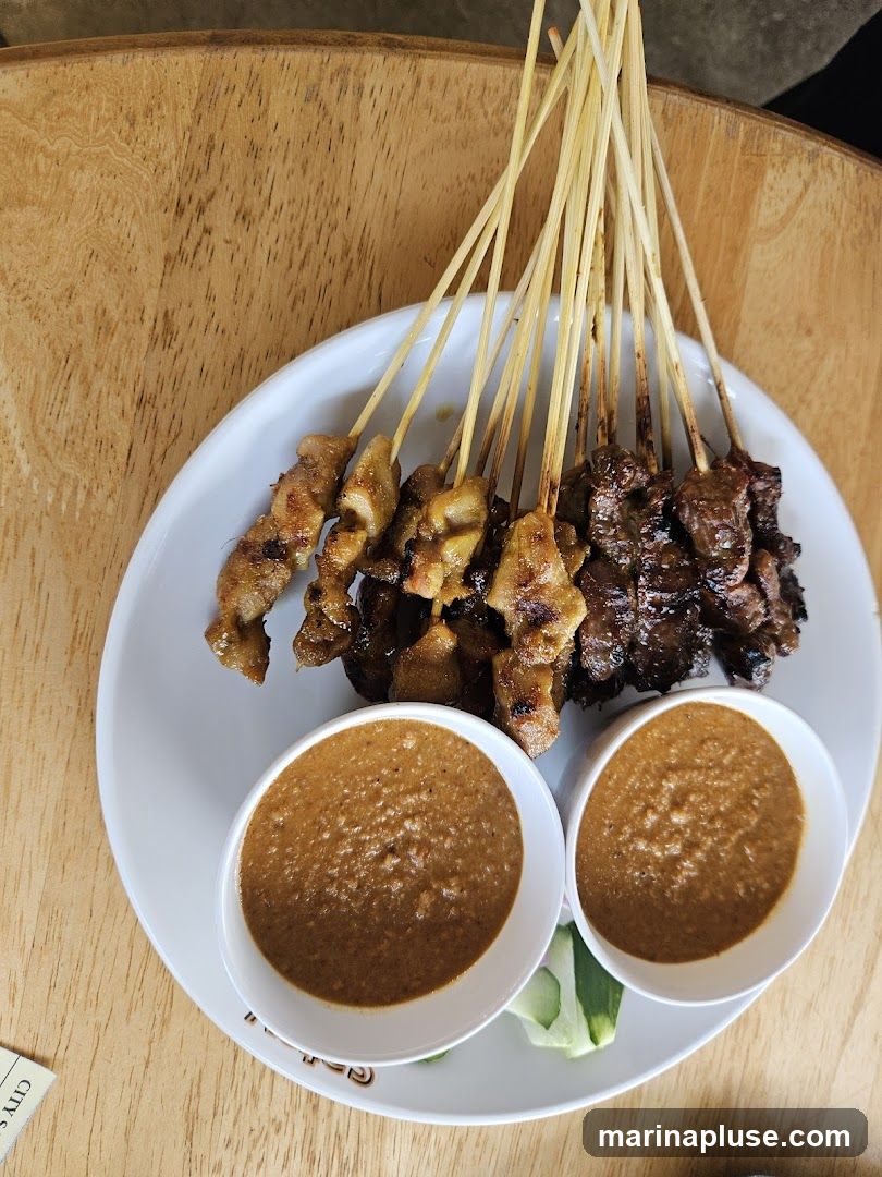 A plate of grilled satay skewers, some appearing to be chicken and others beef, served with two small bowls of peanut sauce and cucumber slices. The skewers are arranged on a white plate on a wooden table.