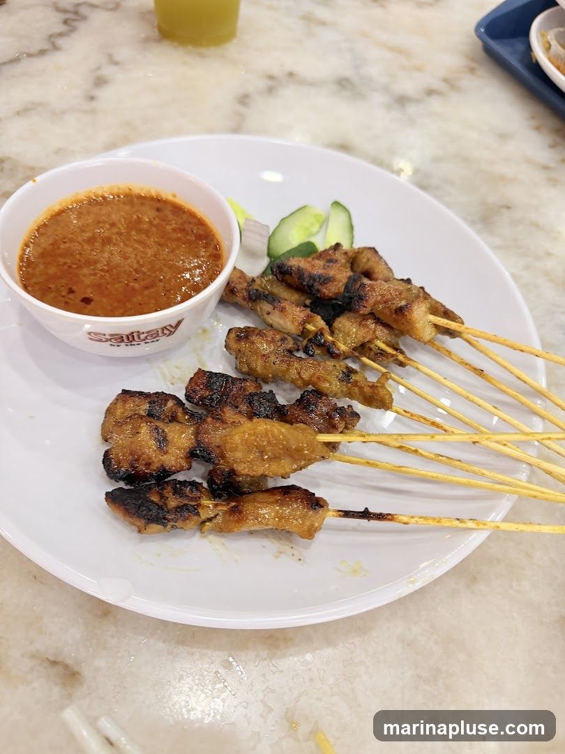 A plate of grilled chicken satay skewers served with a side of peanut sauce and cucumber slices. The satay appears to be well-marinated and grilled to a slight char. The peanut sauce is thick and chunky. The dish is presented on a white plate with a marble-patterned table surface visible. A glass of beverage is partially visible in the background. The logo "Satay by the Bay" is visible on the side of the peanut sauce bowl.