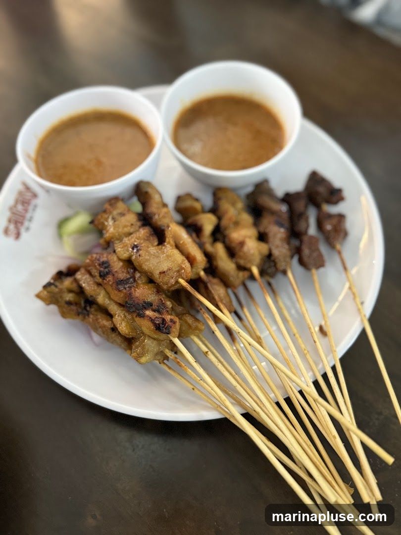 A plate of grilled chicken and beef satay skewers served with two bowls of peanut sauce. The plate has a logo that appears to say "Satay" in red lettering. Sliced cucumbers and red onions are visible under the skewers.