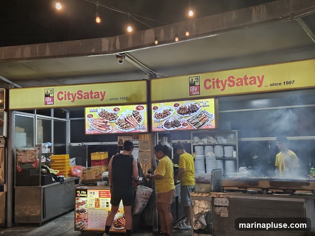 A busy food stall with several people working and ordering. The stall has large yellow signs with the name "CitySatay" and "since 1987". There are illuminated menu boards displaying various satay dishes and other food items. People are seen queuing and interacting with staff. The stall features a grill where satay is being cooked, with smoke rising.