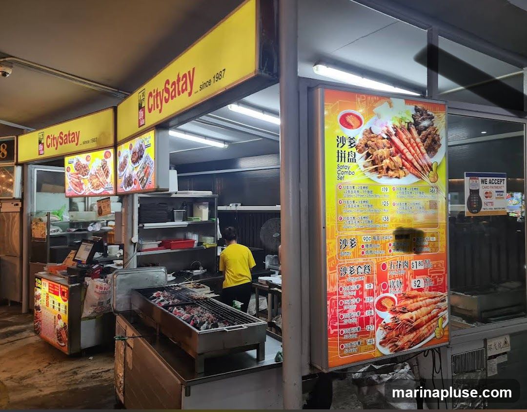 A food stall with a prominent "CitySatay... since 1987" sign. A large illuminated menu board displays various satay combo sets and individual satay options, including prawns and pork belly. A person in a yellow shirt is visible working behind the counter, and a grill with satay skewers is in the foreground.