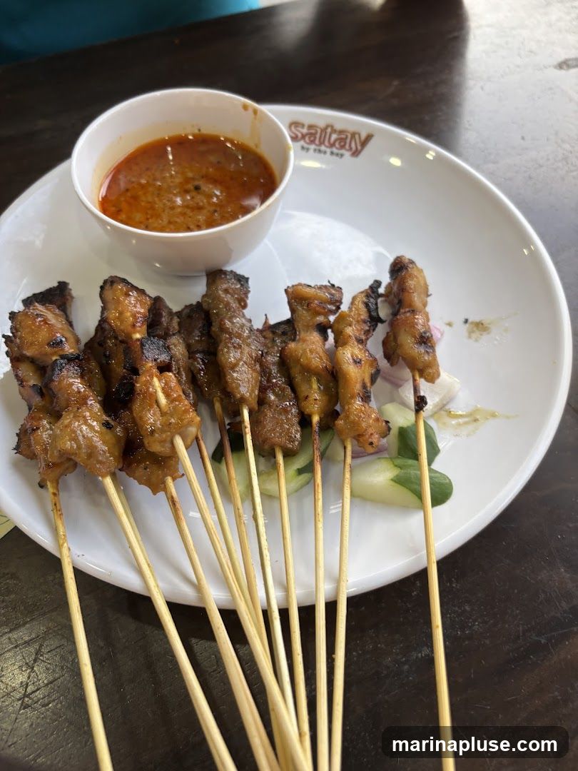 A plate of satay skewers with a side of peanut sauce, cucumber, and red onion. The plate has the logo "Satay by the Bay" printed on it.