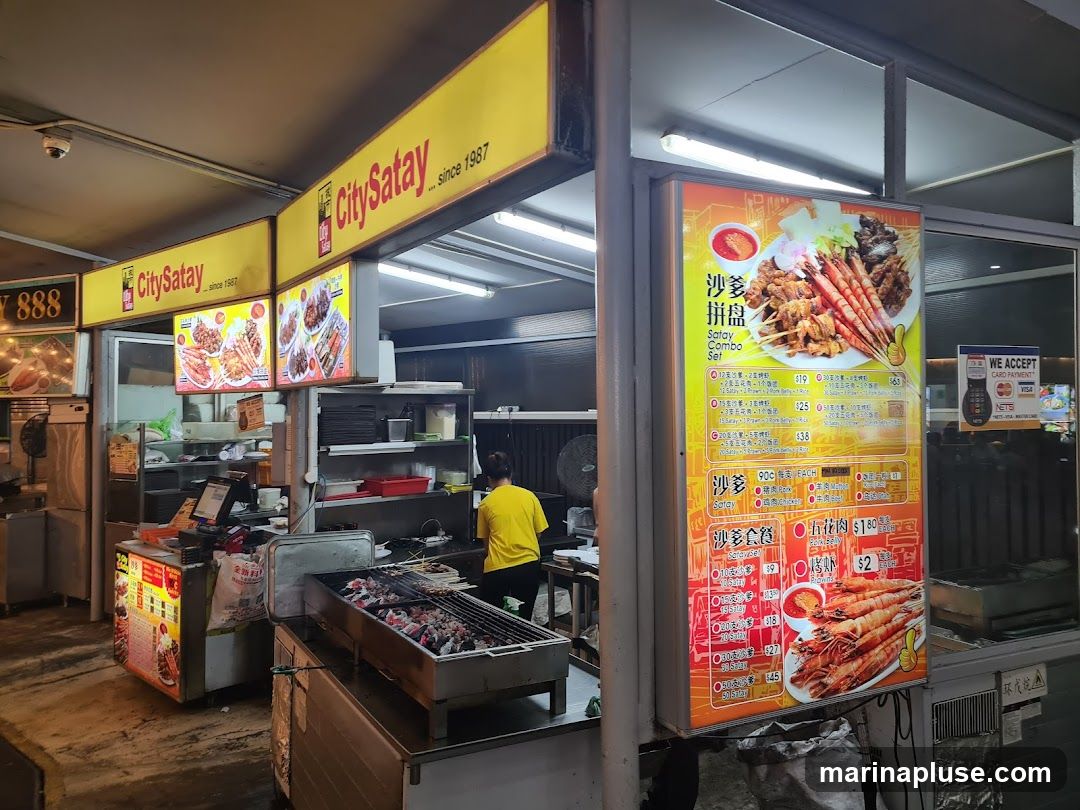 A brightly lit menu board displays various satay sets and individual satay options. The menu features images of satay skewers and prawns. Payment methods accepted are also displayed.