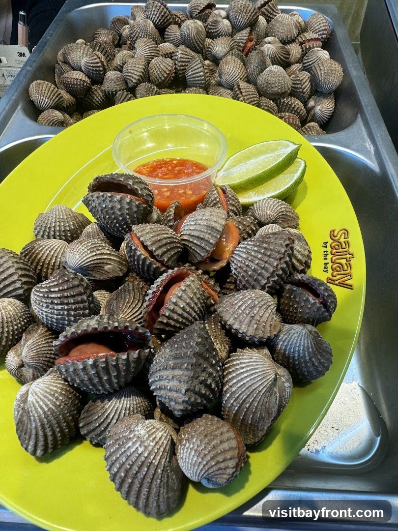 A bright yellow plate filled with cockles, served with a small plastic container of red chili sauce and two lime wedges. In the background, a large metal tray is overflowing with more cockles. The logo 'Salty by the Bay' is visible on the right side of the plate.