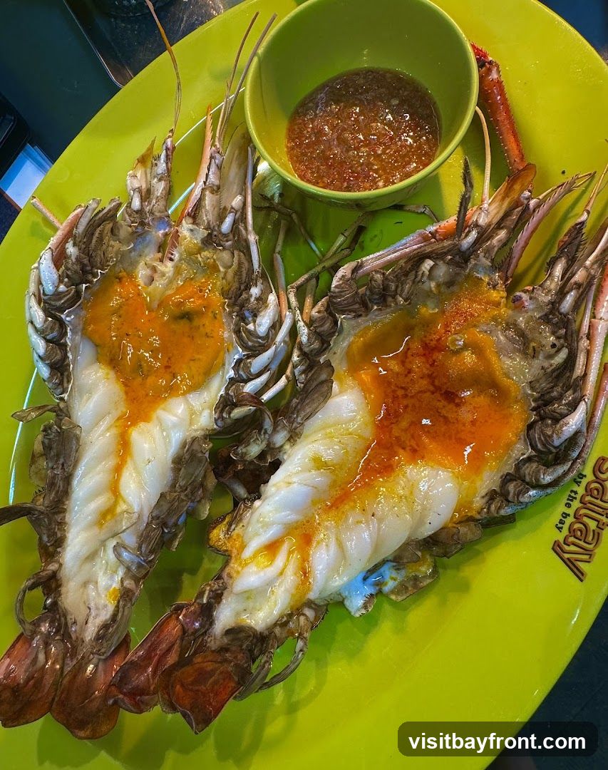 Two large grilled prawns are served on a bright green plate with a small bowl of dipping sauce. The prawns are split lengthwise, revealing their white meat and a rich orange sauce. The plate has a logo that reads "Salay by the bay".