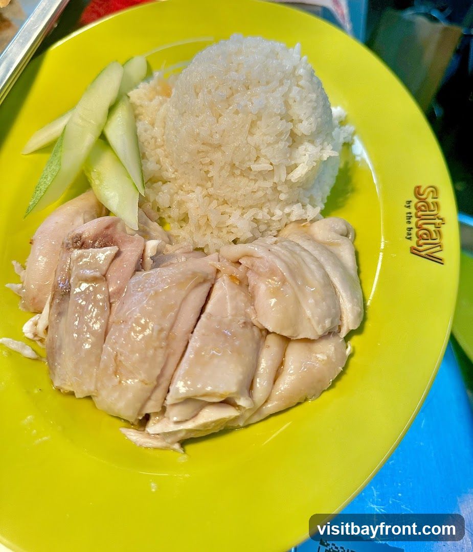 A plate of chicken rice with sliced chicken, rice, and cucumber slices. The plate has a logo that says "Saitay by the bay".