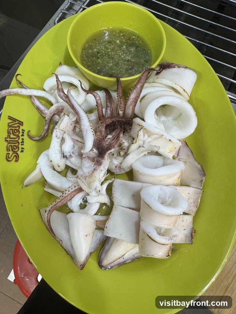A bright green plate holds a serving of grilled squid, including tentacles and sliced rings. A small yellow bowl contains a green dipping sauce. The plate has the text "satay by the bay" printed on it.