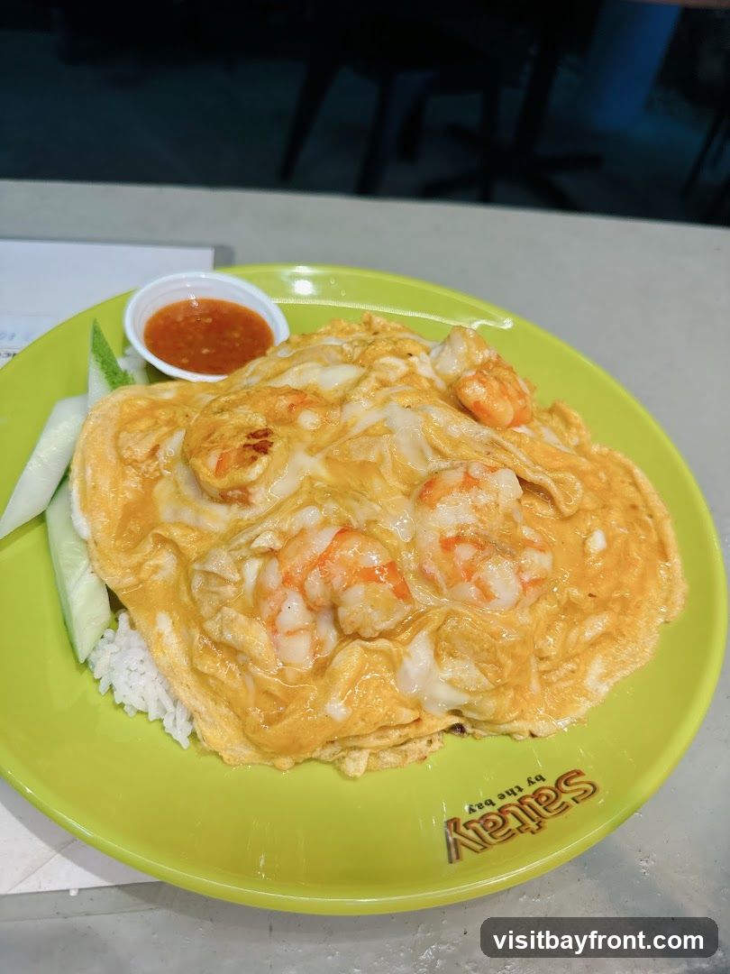 A close-up shot of a plate of rice topped with a fluffy omelette filled with shrimp. Sliced cucumbers and a small dish of chili sauce are served on the side. The plate has the logo "Satay by the Bay" printed on it.