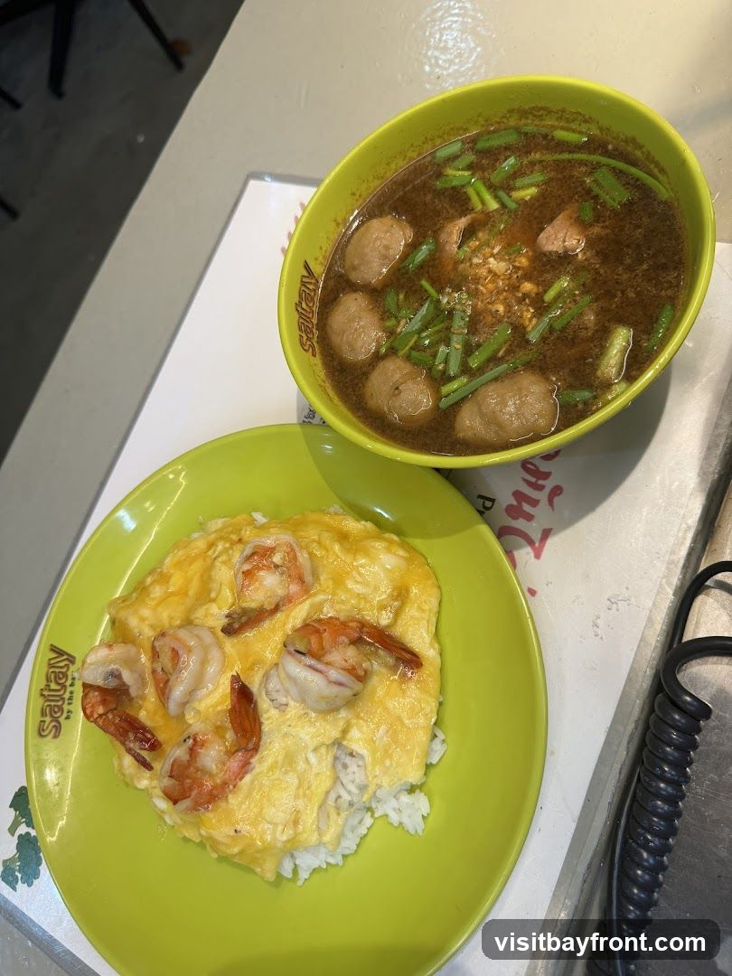 A bright green bowl of noodle soup with meatballs and chopped green onions sits next to a bright green plate of rice topped with an omelet and shrimp. The dishes are on a table with a white placemat that has the logo "Satay by the Bay" printed on it.