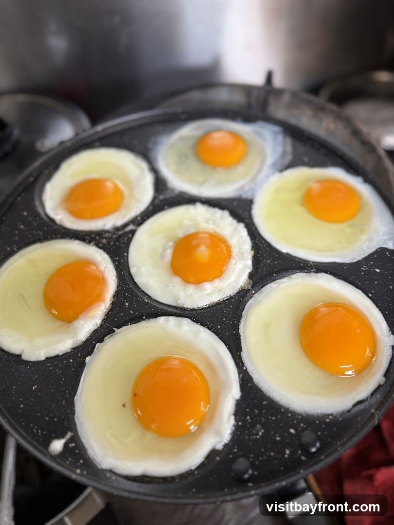 Seven sunny-side-up eggs are cooking in a black non-stick pan with multiple circular indentations. The egg whites are mostly set, and the yolks are bright orange and appear to be runny. The pan is on a stovetop, with a hint of a red cloth visible on the right side.