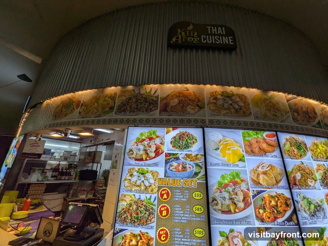 A food stall display featuring various Thai dishes with images and prices. The stall's name, "Kin Aroy Thai Cuisine," is visible above. The display includes dishes like Tom Yum Fried Rice, Oyster Omelette, Mango Salad, Thai Style Fish Cake, and various set meals with prices.