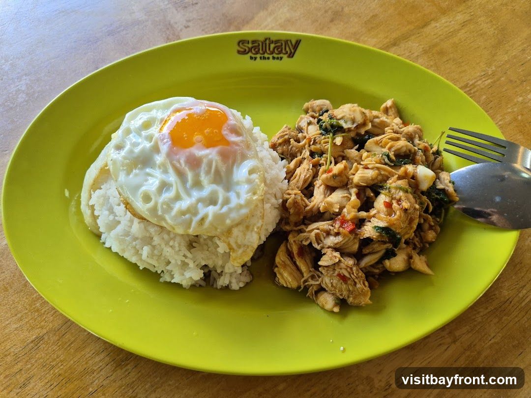 A plate of food with rice, stir-fried chicken with basil and chili, and a fried egg on top of the rice. The plate is lime green and has the logo "satay by the bay" on the rim. A spoon and fork are on the right side of the plate.