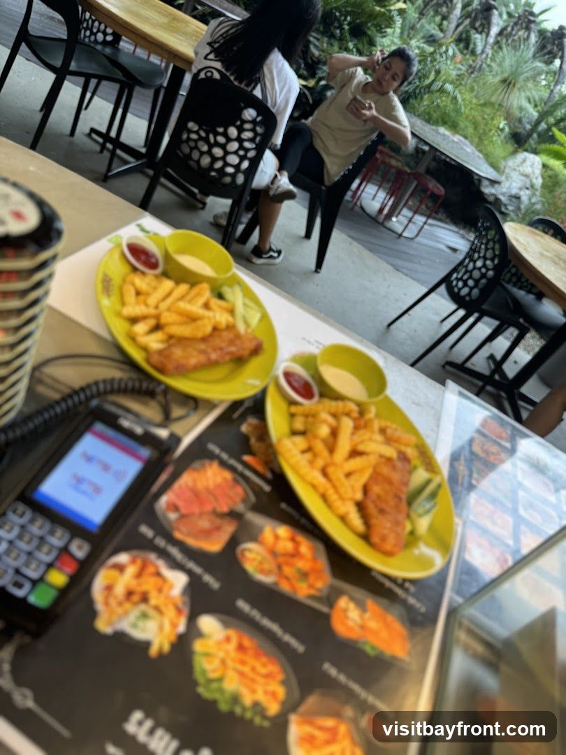 Two plates of fish and chips are served on a table, with a payment terminal and a menu in the foreground. In the background, diners are seated at tables in an outdoor setting with lush greenery.
