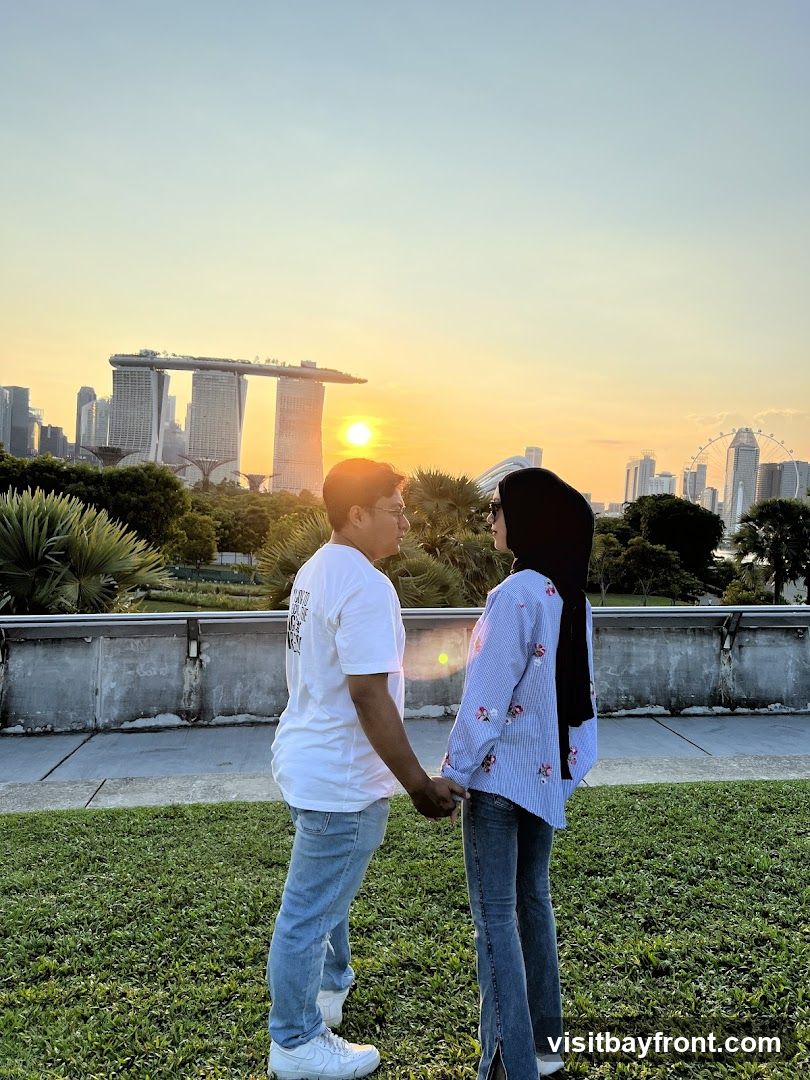 A couple holds hands while facing each other, silhouetted against a vibrant sunset. In the background, the distinctive skyline of a city, including the Marina Bay Sands hotel and a Ferris wheel, is visible. The couple is standing on a grassy area with a concrete barrier behind them.